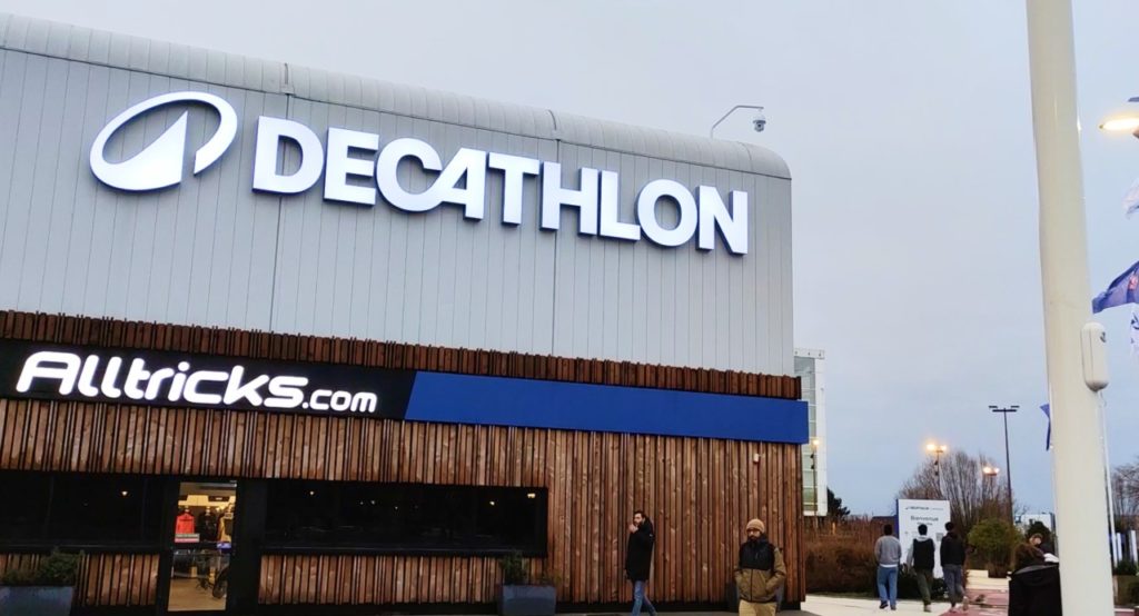 Photo extérieure d'un magasin Decathlon, par temps gris. Sur le côté droit, des drapeaux Decathlon flottent.